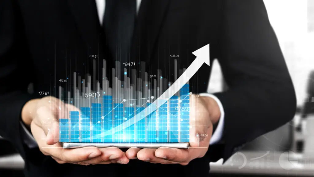 A businessperson in a suit holds a tablet displaying a glowing financial dashboard with blue bar charts, data points, and a large upward arrow, symbolizing business growth, increased revenue, and financial performance.
