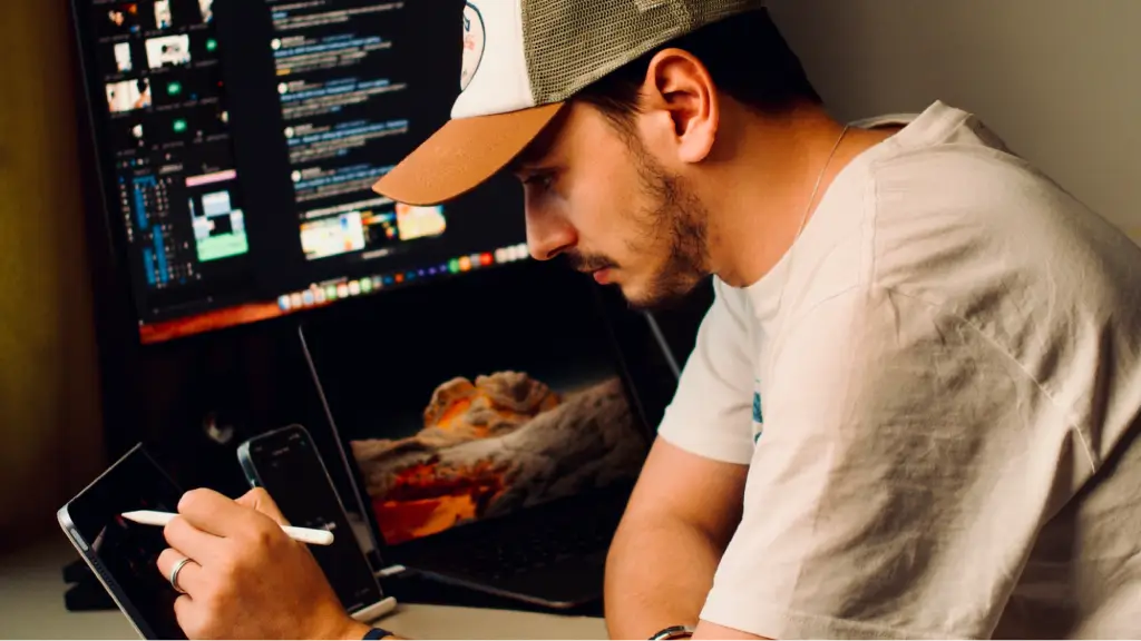 A man wearing a baseball cap sits at his desk, focused on using a stylus on a tablet while surrounded by multiple screens, including an open laptop and a large desktop monitor displaying video editing software.