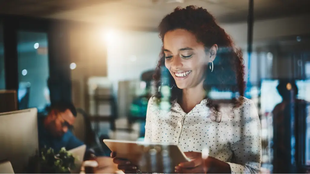 A smiling professional woman using a tablet in a modern office environment, representing productivity, collaboration, digital workflows, and strategic business operations in a technology-driven workplace.