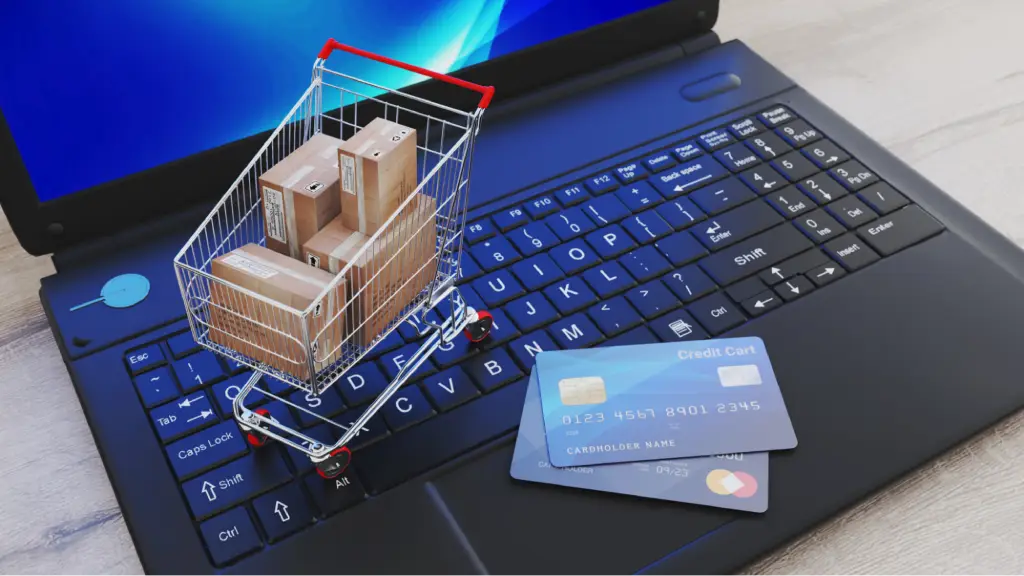 A miniature shopping cart filled with small cardboard boxes resting on a black laptop keyboard next to two simulated credit cards.