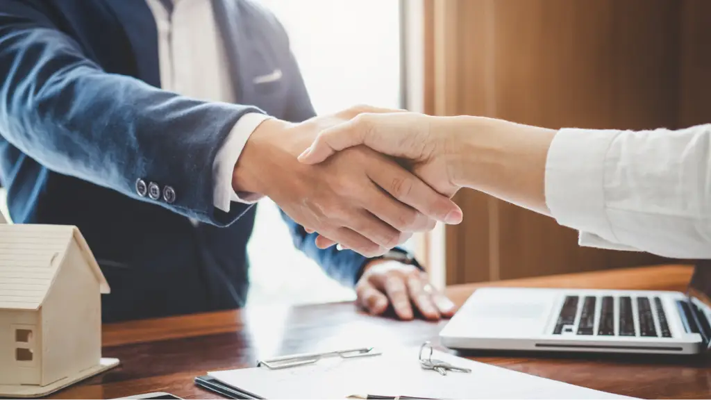 Two people shake hands across a desk with a laptop, documents, and a small house model, representing a successful business agreement, client partnership, or long-term professional relationship.