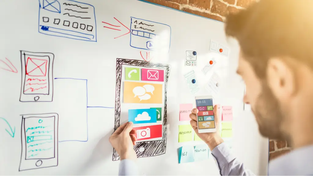 The Rise of Edge AI and Native Cross-Platform Frameworks A person designing a mobile app interface on a whiteboard with sketches of smartphone screens, icons, and user flows, representing mobile app development, user experience design, Edge AI integration, and cross-platform application planning.